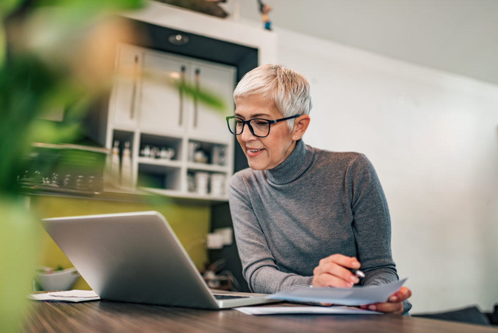 Doing paperwork at home. Senior woman looking at laptop and holding paper document, portrait.